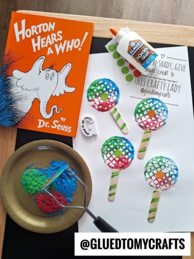 A Horton Hears a Who! Dr. Seuss book, a bottle of Elmer’s glue, finished paper plate crafts resembling Hortons face, craft supplies, paint, and a @GluedToMyCrafts credit label are arranged on a table.