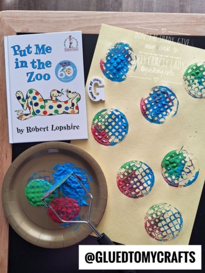 A children’s book titled “Put Me in the Zoo” sits beside a yellow paper with colorful painted circles. A plate holds blue, green, and red paint with a potato masher used as a stamp. ©GluedToMyCrafts.