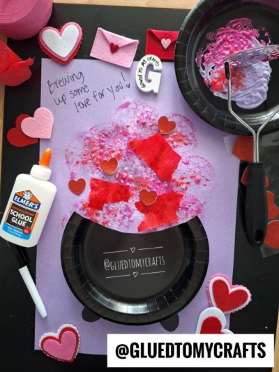 A Valentine’s Day themed craft project with a purple paper, black paper plate, paint roller, glue, felt hearts, and red and pink painted shapes. The text says Brewing up some love for you and @GLUEDTOMYCRAFTS.