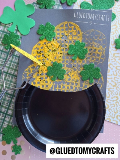 A St. Patrick’s Day craft featuring a black paper plate as a potato masher stamped pot of gold, glittery green shamrocks, and golden mesh spilling out. The background includes green, patterned papers and a label @GLUEDTOMYCRAFTS.