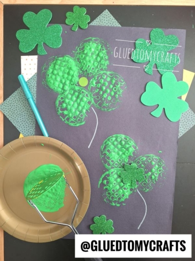 A St. Patrick’s Day craft features green shamrocks, including a potato masher stamped craft, made by stamping bubble wrap with paint on black paper. Glittery shamrock cutouts, a blue paintbrush, green paint, and @GLUEDTOMYCRAFTS are also visible.