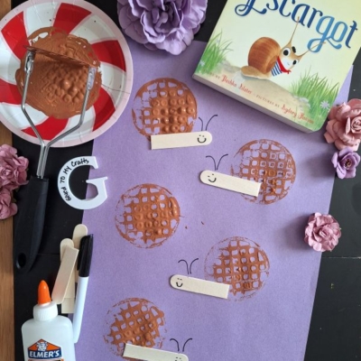 A snail-themed craft setup with a purple paper featuring stamped circles and craft sticks as snail bodies, a Escargot book, glue, paint, a masher tool, and flowers. @GLUEDTOMYCRAFTS label at the bottom.