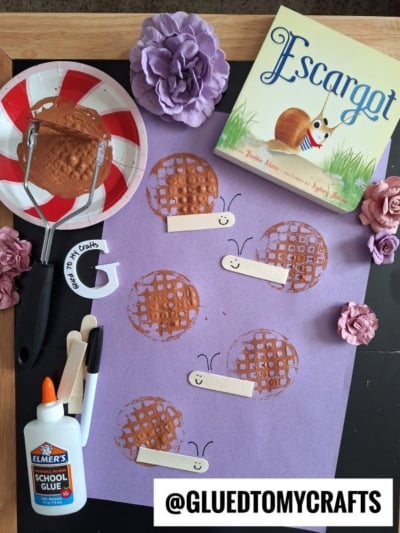A snail-themed craft setup with a purple paper featuring stamped circles and craft sticks as snail bodies, a Escargot book, glue, paint, a masher tool, and flowers. @GLUEDTOMYCRAFTS label at the bottom.