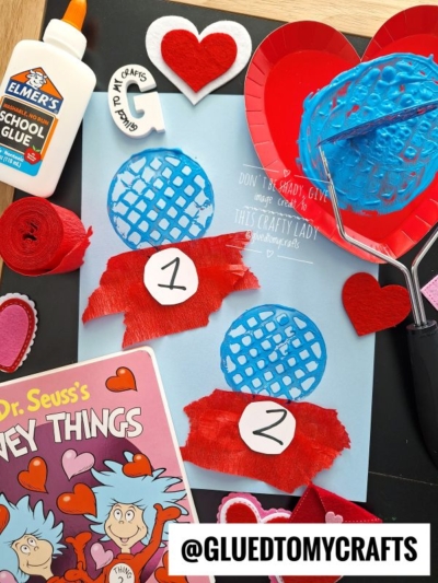 A Dr. Seuss-inspired craft scene with two blue fishbowls labeled 1 and 2 on blue paper, red felt hearts, red crepe paper, glue, and a Cat in the Hat book surrounded by crafting supplies.