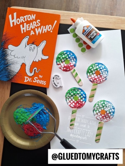 A Dr. Seuss book, a bottle of glue, paint, and a paper with four painted lollipops made from mesh and craft sticks are arranged on a table. A gold plate holds painted mesh, and @GLUEDTOMYCRAFTS is written at the bottom.