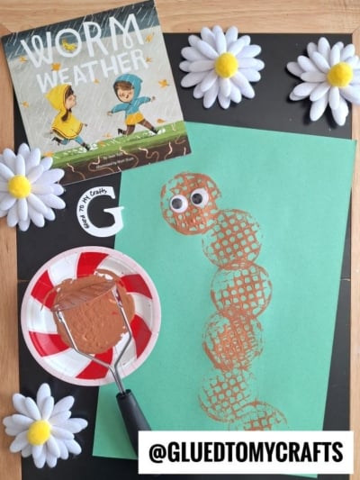 A childrens craft display shows a green paper with a stamped brown worm shape, googly eyes, and a book titled Worm Weather. White fabric daisies, a paint roller, and a red-striped plate with paint are included.