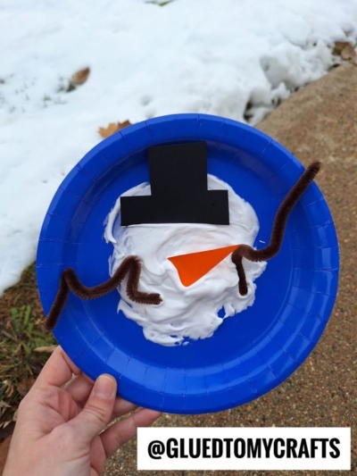 A blue paper plate decorated as a snowman face with puffy paint crafts like shaving cream, a black paper hat, orange paper nose, and brown pipe cleaner arms. Snow covers the ground in the background. Text reads @GLUEDTOMYCRAFTS.
