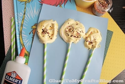Three yellow, swirled, sprinkle-covered treats on striped sticks rest on blue paper, surrounded by craft supplies, glitter, and a Dr. Seuss book.