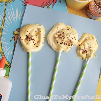 Three yellow, swirled, sprinkle-covered treats on striped sticks rest on blue paper, surrounded by craft supplies, glitter, and a Dr. Seuss book.