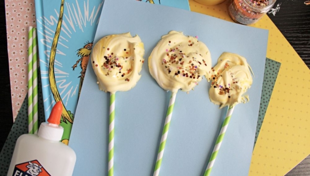 Three yellow, swirled, sprinkle-covered treats on striped sticks rest on blue paper, surrounded by craft supplies, glitter, and a Dr. Seuss book.