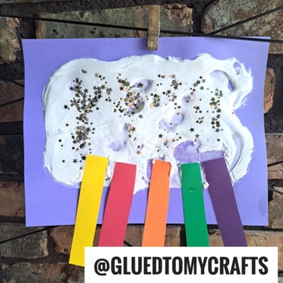 A children’s craft with stars and glitter on white glue spread over purple paper, with red, orange, yellow, green, and purple paper strips attached at the bottom. The craft is clipped to a wire.