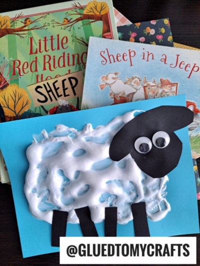 A sheep craft made with shaving cream and paper sits on a blue card in front of childrens books, including Little Red Riding Hood and Sheep in a Jeep. The card has googly eyes and black paper legs and face.