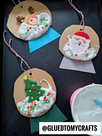 Three handmade Christmas ornaments on brown paper circles—reindeer, Santa, and a Christmas tree—each decorated with puffy paint crafts, white glue, colorful beads, and string for hanging. A bowl of glue is nearby. @GLUEDTOMYCRAFTS is at the bottom.