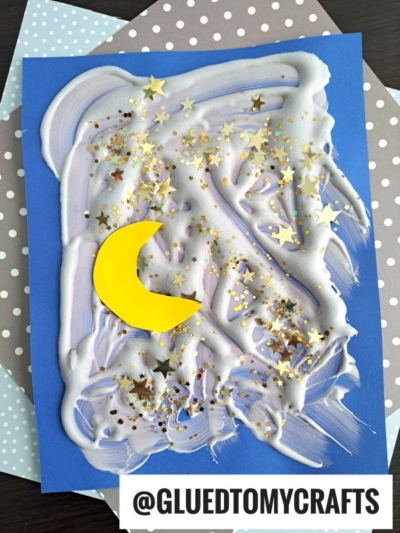 Children’s artwork of a night sky made with white and purple puffy paint crafts, gold star-shaped glitter, and a yellow crescent moon on blue paper. Polka dot papers are underneath. @GLUEDTOMYCRAFTS is written at the bottom.