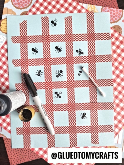 A charming insect craft idea featuring a red and white gingham paper, blue squares, and ant drawings. Nearby are a black marker, black paint, and a cotton swab. @GLUEDTOMYCRAFTS is noted at the bottom.