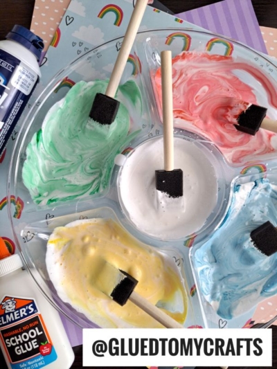 A palette with five sections holds colorful homemade paints in green, white, pink, yellow, and blue, each with a foam brush. Bottles of Elmer’s glue and blue acrylic paint are nearby. Background has rainbow-patterned paper.