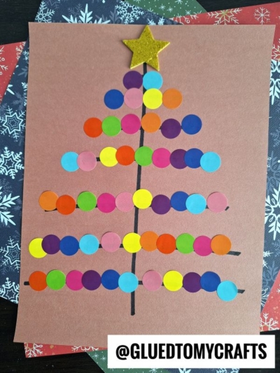 A Christmas tree made from colorful paper dots arranged in rows on brown paper, with a yellow star at the top and a black tree trunk beneath. Coding label crafts add a playful touch, while festive snowflake pattern paper decorates the background.
