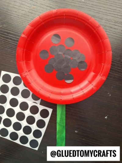 A red paper plate, decorated with black dot stickers to resemble a watermelon, features a green craft stick “stem” at the bottom—perfect for coding label crafts. Nearby, a sheet of unused black dot stickers rests on the dark surface.