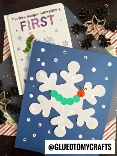 A blue card with a white snowflake decorated with green circles and an orange face to resemble a caterpillar, inspired by The Very Hungry Caterpillar book, surrounded by snowflake ornaments—perfect for fun coding label crafts.