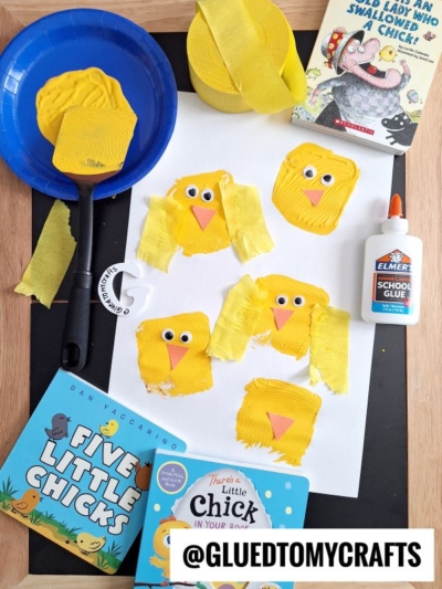A children’s craft scene with yellow chick faces made from painted cardboard squares, googly eyes, and paper beaks on white paper. Surrounding the craft are books, yellow ribbon, glue, and a blue plate.