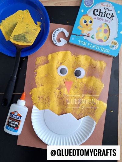 A paper plate chick craft made with yellow paint, large googly eyes, an orange beak, glue, and a brown background. Paint and a book titled Theres a Little Chick in Your Book are nearby.