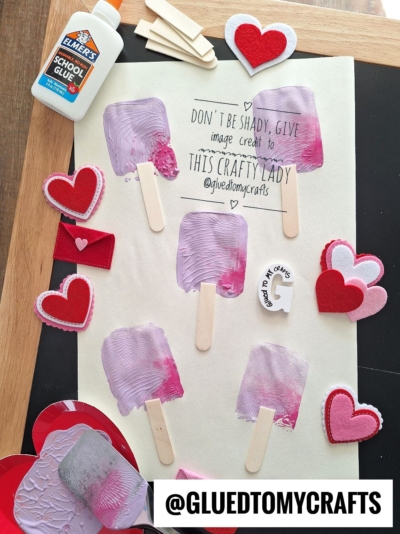 Art project with popsicle stick crafts painted in purple and red, surrounded by felt hearts, a glue bottle, and a sign reading “Don’t be shady, give image credit to this crafty lady @gluedtomycrafts.”.