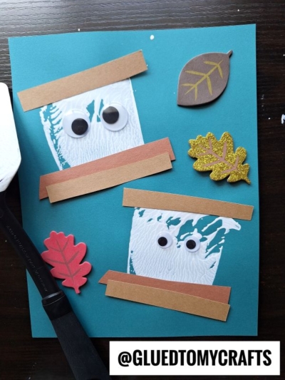 Two white ghost crafts with googly eyes and brown paper strips on blue paper, decorated with three foam leaves; a spatula is on the left and @GLUEDTOMYCRAFTS appears at the bottom.