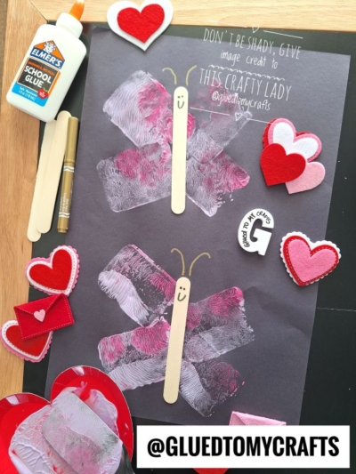A childrens craft project with two butterflies made from white and pink handprints and craft sticks on black paper, surrounded by felt hearts, a gold marker, glue, craft sticks, and an @ handle tag.