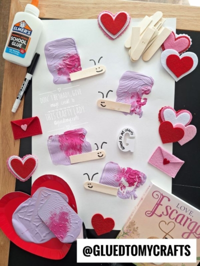 A flat lay of Valentine-themed crafts: popsicle stick caterpillars painted purple with smiley faces, red and pink heart decorations, envelopes, a glue bottle, marker, and a book, arranged on a white surface.