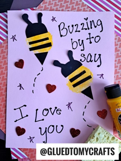 A handmade card with two bee shapes, “Buzzing by to say I love you,” red glitter hearts, and a yellow paint tube—perfect for Sponge Painting Ideas for Kids. The background features layered pink and patterned paper.