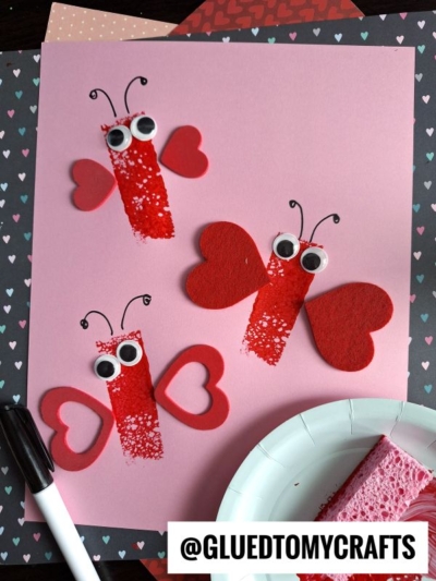 Three butterfly crafts made from red paint, googly eyes, and red paper hearts are displayed on pink paper—perfect for Sponge Painting Ideas for Kids. A black marker, white plate with a pink sponge, and heart-patterned papers complete the scene.