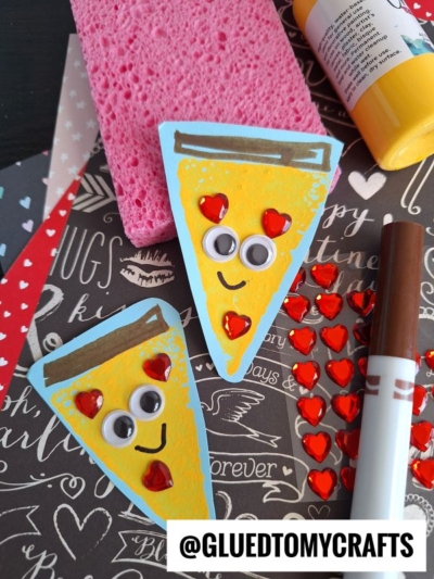 Two pizza slice crafts with googly eyes, red heart stickers as pepperoni, and smiles sit on a patterned surface—perfect Sponge Painting Ideas for Kids—alongside a pink sponge, yellow paint bottle, brown marker, and extra heart stickers.