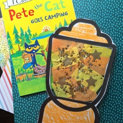 A Pete the Cat Goes Camping book lies next to a handmade lantern craft decorated with orange, yellow, and gold tissue paper, glitter, and star shapes. Both items rest on colorful, patterned paper backgrounds.