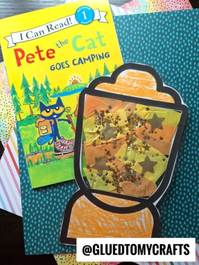 A Pete the Cat Goes Camping book lies next to a handmade lantern craft decorated with orange, yellow, and gold tissue paper, glitter, and star shapes. Both items rest on colorful, patterned paper backgrounds.