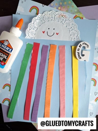 A paper craft shows a smiling cloud made from a white doily and paper, with colorful paper strips as rainbows, glue, and a paintbrush on a blue background with rainbow-patterned papers. @GLUEDTOMYCRAFTS is at the bottom.