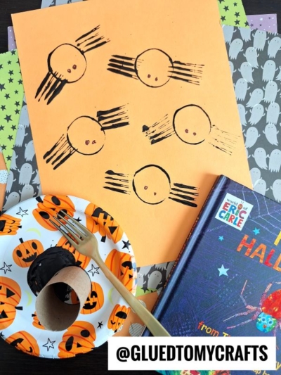 A Halloween craft scene featuring INSECT CRAFT IDEAS: an orange paper with black-painted circles and lines, made with a cardboard tube and black paint. Surrounding items include a pumpkin plate, fork, Halloween book, and ghost-patterned paper.