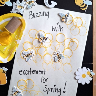 A craft sheet with yellow paint honeycomb circles, bee stickers, and the handwritten message “Buzzing with excitement for Spring!” Bees, flowers, and paint are arranged on a black background. @GLUEDTOMYCRAFTS is at the bottom.