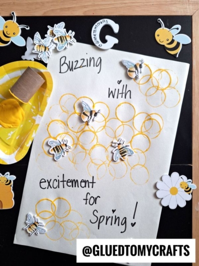 A craft sheet with yellow paint honeycomb circles, bee stickers, and the handwritten message “Buzzing with excitement for Spring!” Bees, flowers, and paint are arranged on a black background. @GLUEDTOMYCRAFTS is at the bottom.