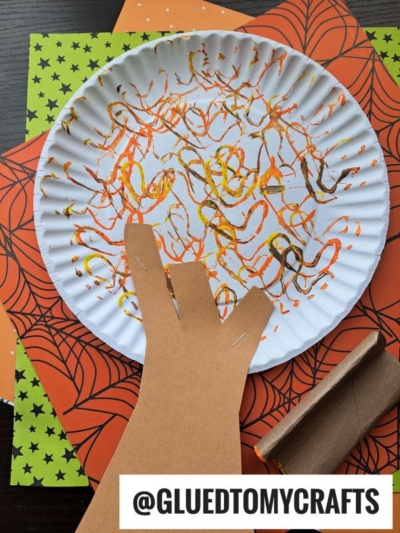 A paper plate decorated with orange, yellow, and black paint squiggles sits on top of orange and spiderweb-patterned paper. A brown paper hand and a cardboard tube are attached, making it perfect for FALL CRAFTS FOR KIDS. @GLUEDTOMYCRAFTS.