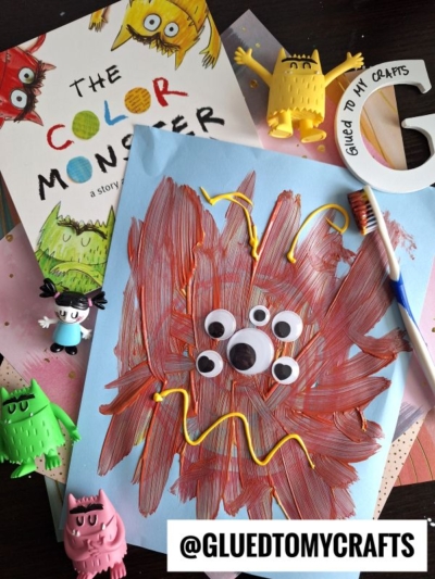 A children’s craft inspired by The Color Monster book, perfect for back to school story time, features a monster made from brown paint, googly eyes, and yellow squiggles on blue paper, surrounded by colorful monster toys and craft supplies.