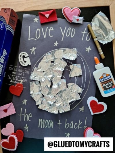 A craft project on black paper features the words Love you to the moon & back! with a moon made of crumpled foil pieces, surrounded by white stars, felt hearts, Elmer’s glue, and a gold pen. @GLUEDTOMYCRAFTS is displayed at the bottom.