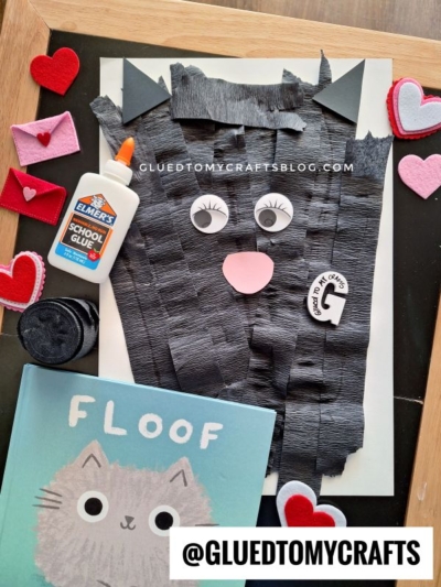 A handmade black cat craft with fringed paper, pink nose, and googly eyes is on a chalkboard, surrounded by a glue bottle, felt hearts and envelopes, and a “Floof” cat book. The username @GLUEDTOMYCRAFTS is visible.