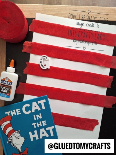 A Cat in the Hat craft setup with red crepe paper strips on white paper, a glue bottle, a Cat in the Hat book, and a small paper circle, on a black background. @GLUEDTOMYCRAFTS is displayed at the bottom.