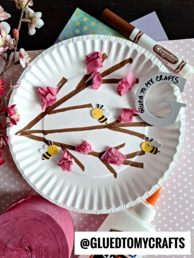A paper plate craft shows a tree branch drawn with brown marker, adorned with pink tissue flowers and smiling bee stickers. Craft supplies and floral decor surround it. The handle @GLUEDTOMYCRAFTS appears on the image.