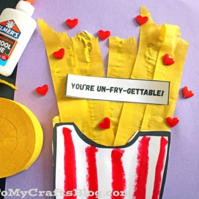 A craft project with yellow crepe paper fries in a red-striped paper container, a label reading Youre UN-FRY-GETTABLE!, red heart decorations, Elmers glue, and a roll of yellow crepe paper on a purple background.
