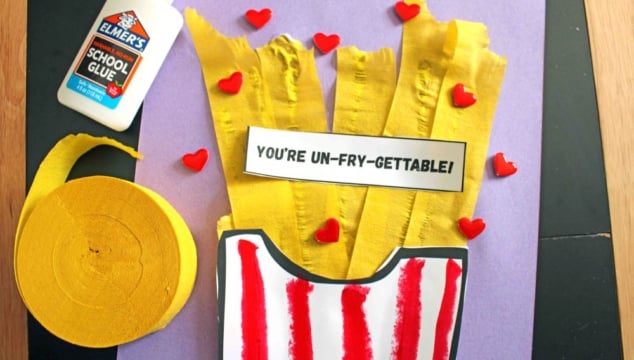 A craft project with yellow crepe paper fries in a red-striped paper container, a label reading Youre UN-FRY-GETTABLE!, red heart decorations, Elmers glue, and a roll of yellow crepe paper on a purple background.