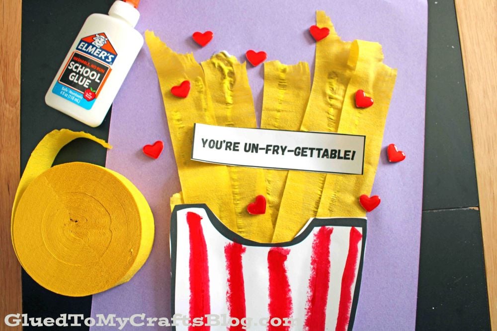 A craft project with yellow crepe paper fries in a red-striped paper container, a label reading Youre UN-FRY-GETTABLE!, red heart decorations, Elmers glue, and a roll of yellow crepe paper on a purple background.