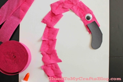 A craft project of a torn crepe paper flamingo made with pink strips, a large googly eye, and black paper for the beak on a white background. A roll of pink crepe paper and a glue bottle are nearby.