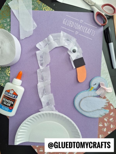A purple background displays a swan craft made from a paper plate, torn crepe paper strips, an orange beak, and a googly eye. Craft supplies, glue, scissors, and a drawing of a sleeping swan are also visible.