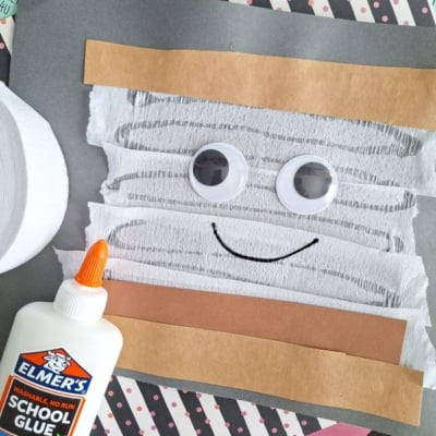 A mummy craft made from white crepe paper strips, googly eyes, and brown paper on a gray background. Elmer’s glue bottle and stacked craft papers are nearby. @GLUEDTOMYCRAFTS is written at the bottom.